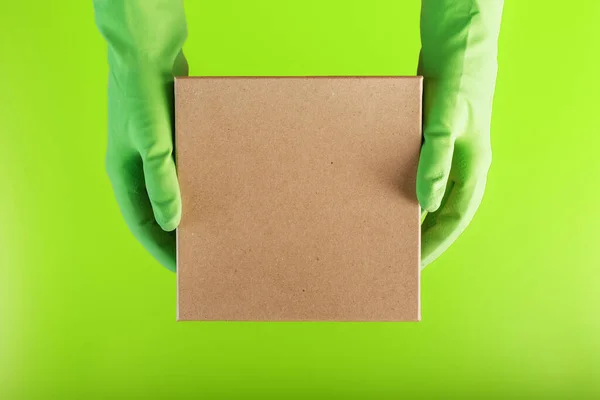 A square box in the hands with green rubber gloves on a green background. Cardboard square mailbox with virus protection.