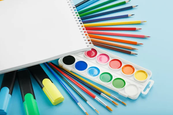 An album for drawing and creativity for school with stationery, a palette of colored paints, markers, brushes and pencils. Free space for text on a blue background. The view from the top