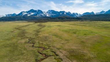 İçinden nehir akan geniş, açık bir alan. Gökyüzü açık ve arka plandaki dağlar karla kaplı. Sahne huzurlu ve huzurlu, nehirle birlikte