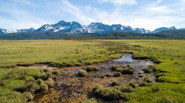 İçinden akarsu akan geniş, açık bir alan. Gökyüzü açık ve arka plandaki dağlar karla kaplı. Sahne huzurlu ve sakin, suyla birlikte.