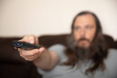 Uzun saçlı bir adam uzaktan kumanda tutuyor. O bir kanepede oturuyor. Uzaktan kumanda siyah ve kırmızı düğmesi var.