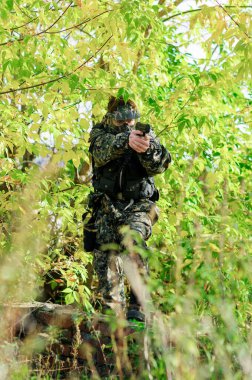 Elinde bir tabanca, kamuflaj üniforması, koruyucu bir maske ve ağaçların arasındaki eğitim alanında taktik bir tatbikatta gözlük takan bir asker.