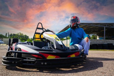 Kasklı genç bir yarışçı gün batımında yarış kartının yanında oturur.