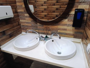 Modern banyo içi iki beyaz seramik lavabo, bir musluk ve yuvarlak ayna, yanında bir el kurutma makinesi, bir kafede ya da alışveriş merkezinde bir tuvalet.