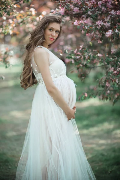Krásná těhotná žena má na sobě bílé šaty dívka pózuje v růžové jablko zahrady, letní čas — Stock fotografie