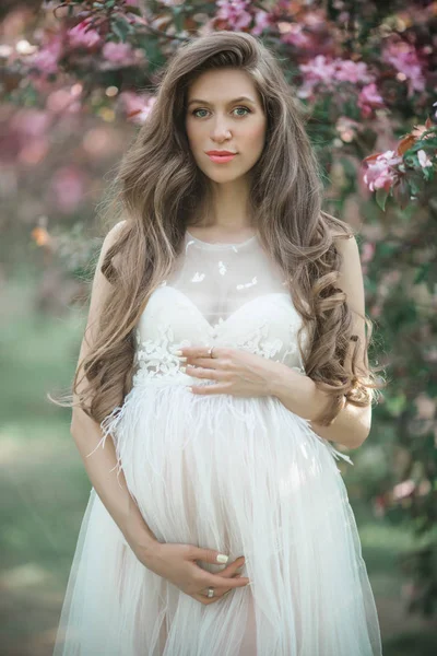 Schwangere hübsche Frau trägt weißes Modekleid posiert im rosa blühenden Apfelgarten, Sommerzeit — Stockfoto