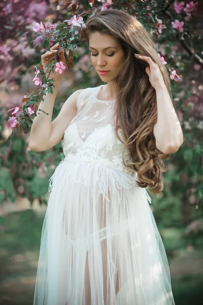 Mujer bonita embarazada está usando vestido de moda blanca posando en el jardín de manzanas de flor rosa, hora de verano — Foto de Stock