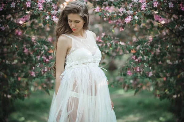 Schwangere hübsche Frau trägt weißes Modekleid posiert im rosa blühenden Apfelgarten, Sommerzeit — Stockfoto