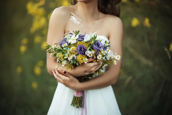 Hermosa novia joven con ramo de flores silvestres en el campo sobre las luces del atardecer — Foto de Stock