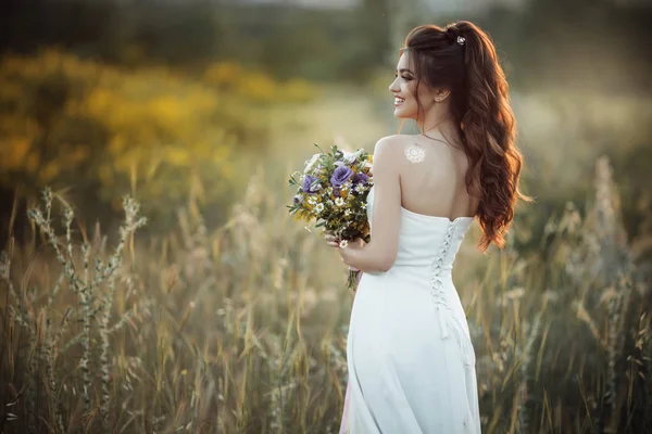 Mooie jonge bruid met boeket van wilde bloemen in veld over zonsondergang lichten — Stockfoto