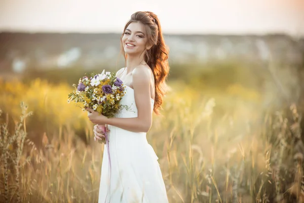 Hermosa novia joven con ramo de moda de flores silvestres en el campo sobre las luces del atardecer — Foto de Stock