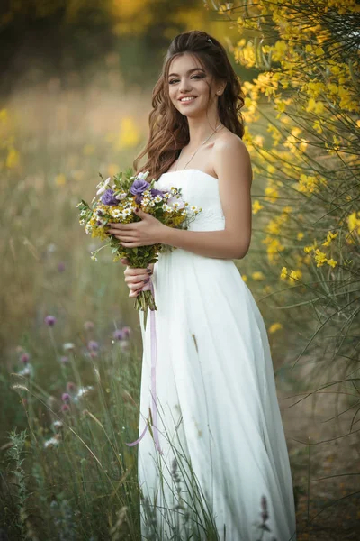 Beautiful lovely bride is wearing white fashion dress holding bouquet of wildflowers in yellow field, nature wedding concept — Stock Photo, Image