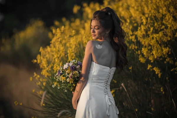 Beautiful lovely bride is wearing white fashion dress holding bouquet of wildflowers in yellow field, nature wedding concept — Stock Photo, Image