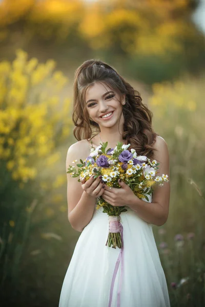 Beautiful smiling bride is wearing white fashion dress holding bouquet of wildflowers in yellow field, nature wedding concept — Stock Photo, Image