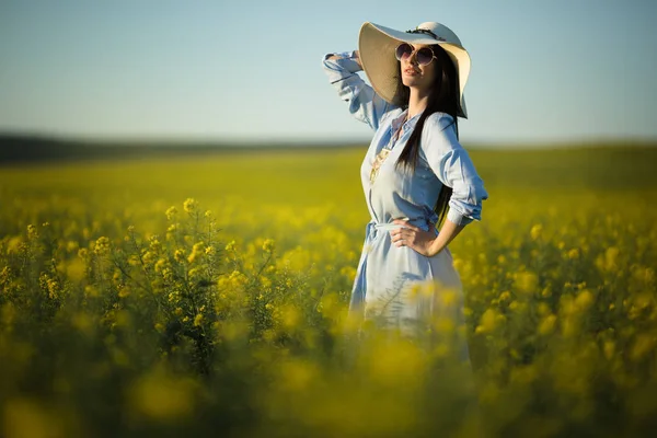 Feliz joven mujer está usando sombrero de moda con ramo de flores silvestres en el campo amarillo en las luces del atardecer — Foto de Stock