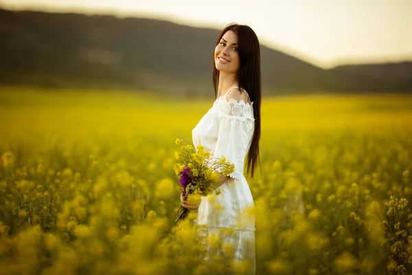 Mujer joven feliz con ramo de flores silvestres en el campo amarillo en las luces del atardecer, hora de verano — Foto de Stock