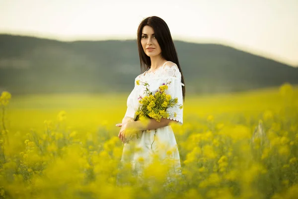 Mujer bonita con ramo de flores silvestres en el campo amarillo en las luces del atardecer, hora de verano — Foto de Stock