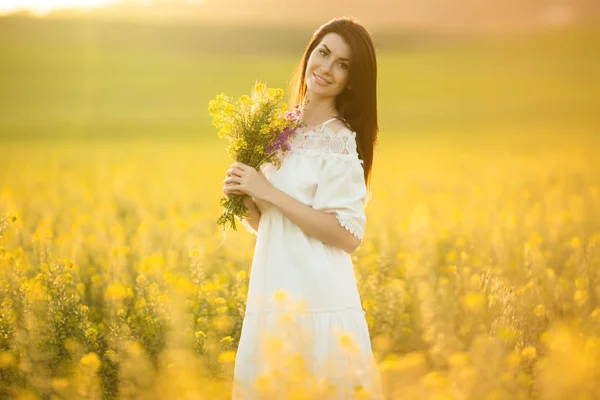 Mulher bonita com buquê de flores silvestres no campo amarelo em luzes de pôr do sol, hora de verão — Fotografia de Stock