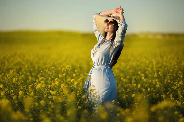 Feliz joven mujer está usando sombrero de moda con ramo de flores silvestres en el campo amarillo en las luces del atardecer — Foto de Stock