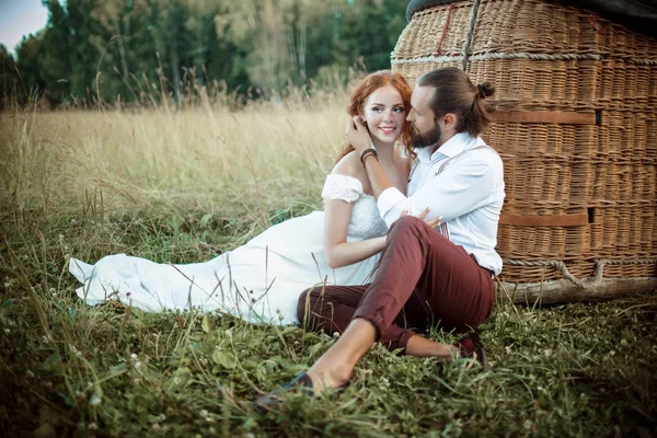 Verheugd huwelijksfeest paar zitten in de buurt van luchtballon mand in het zonnige veld. — Stockfoto