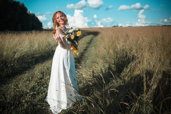 Vackra brud med fräknar på hennes ansikte bär vit mode klänning i fältet med brudbukett av vildblommor — Stockfoto