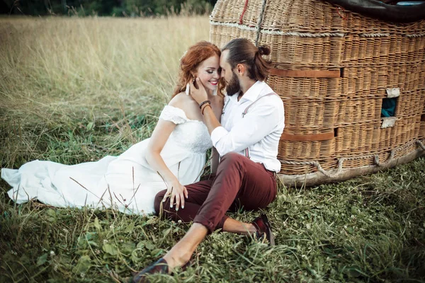 Heureux couple de mariage sont assis près du panier aéroballon dans le champ ensoleillé . — Photo