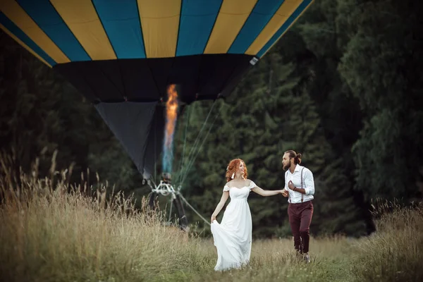 Feliz casamento lindo casal noiva e noivo estão andando perto de cesta de balão de ar no campo . — Fotografia de Stock