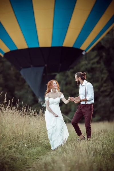 Feliz casamento lindo casal noiva e noivo estão andando perto de cesta de balão de ar no campo . — Fotografia de Stock
