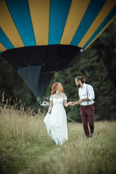 Feliz casamento lindo casal noiva e noivo estão andando perto de cesta de balão de ar no campo . — Fotografia de Stock