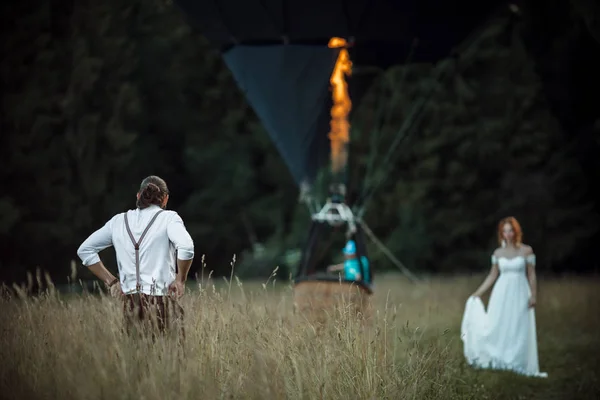 Feliz casal encantador noiva e noivo estão andando perto de cesta de balão de ar no campo, casamento e conceito de aventura — Fotografia de Stock