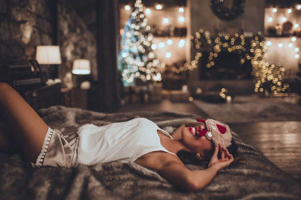 Όμορφη γυναίκα της Ασίας είναι ξαπλωμένη στο κρεβάτι και φορώντας μάσκα ύπνου στο σπίτι κοντά στο χριστουγεννιάτικο δέντρο στο άνετο εσωτερικό. Εσωτερικό με χριστουγεννιάτικα στολίδια. — Φωτογραφία Αρχείου