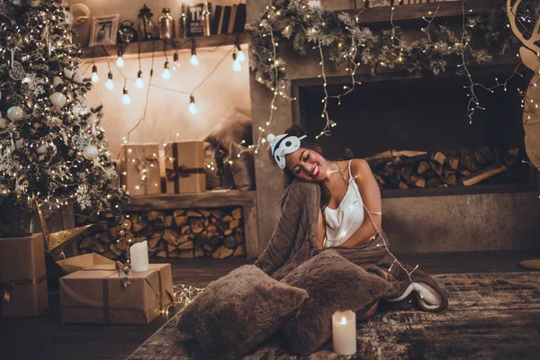 Όμορφη ασιατική γυναίκα φοράει στον ύπνο μάσκα και πιτζάμες στο σπίτι κοντά στο χριστουγεννιάτικο δέντρο και τζάκι στο άνετο εσωτερικό. Εσωτερικό με χριστουγεννιάτικα στολίδια. — Φωτογραφία Αρχείου