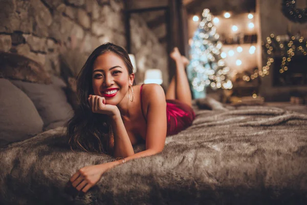 Mulher asiática bonita está sentada na cama em casa perto da árvore de Natal no interior acolhedor. Interior com decorações de Natal . — Fotografia de Stock