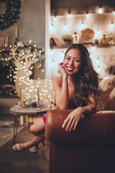 Ung Vacker asiatisk kvinna är sexig röd klänning sitter hemma nära julgran i mysig interiör. Interiör med juldekorationer. — Stockfoto