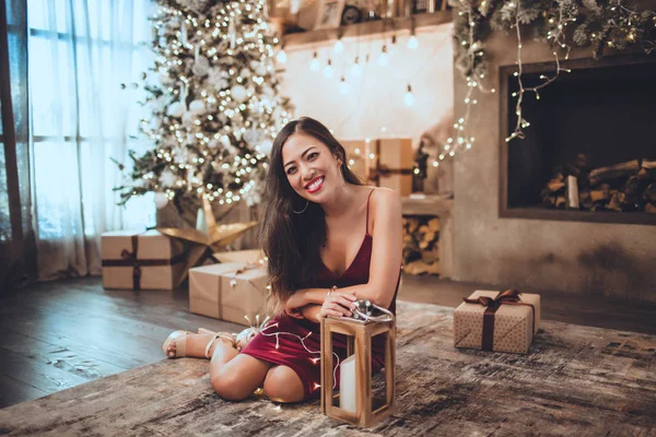 Ung Vacker asiatisk tjej är fashion röd klänning sitter hemma nära julgran i mysig interiör. Interiör med juldekorationer. — Stockfoto