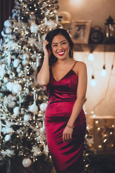 美しいアジア人の少女が赤いファッション ドレス家で居心地のよいインテリアでクリスマス ツリーのそばに座っています。クリスマスの装飾とインテリア. — ストック写真