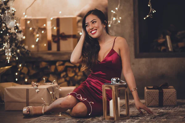 Νέοι όμορφο κορίτσι της Ασίας είναι μόδα κόκκινο φόρεμα κάθεται στο σπίτι κοντά στο χριστουγεννιάτικο δέντρο στο άνετο εσωτερικό. Εσωτερικό με χριστουγεννιάτικα στολίδια. — Φωτογραφία Αρχείου