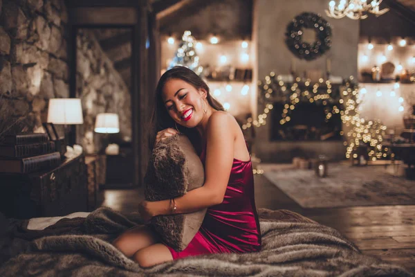 Vacker asiatisk kvinna sitter i sängen hemma nära julgran i mysig interiör. Interiör med juldekorationer. — Stockfoto