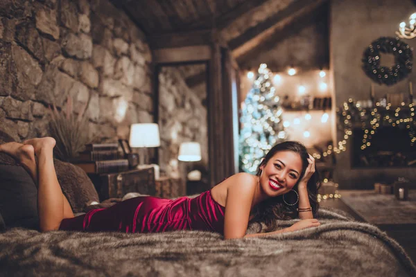 Vackra glada asiatisk kvinna sitter i sängen hemma nära julgran i mysig interiör. Interiör med juldekorationer. — Stockfoto
