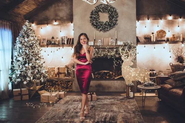 Νέοι όμορφο κορίτσι της Ασίας είναι μόδα κόκκινο φόρεμα κάθεται στο σπίτι κοντά στο χριστουγεννιάτικο δέντρο στο άνετο εσωτερικό. Εσωτερικό με χριστουγεννιάτικα στολίδια. — Φωτογραφία Αρχείου