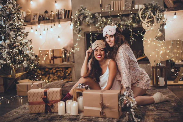 Δύο φίλοι ευτυχισμένη κορίτσια φορούν μάσκα ύπνου στο σπίτι κοντά στο χριστουγεννιάτικο δέντρο στο άνετο εσωτερικό. Εσωτερικό με χριστουγεννιάτικα στολίδια. — Φωτογραφία Αρχείου