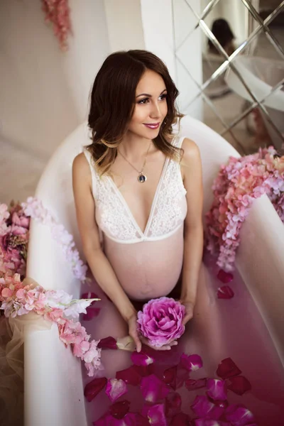 美しい妊娠中の女性は紫水とバラの花びらのバスタブに横たわって白のランジェリーを着ています。 — ストック写真