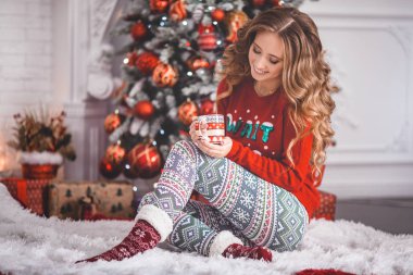 Güzel genç kız hediye kutusu ile Noel ağacının yanında oturan ev rahat xmas kıyafetlerini giyiyor