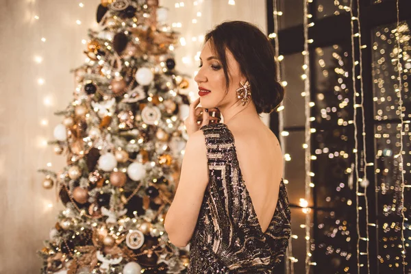 Mulher bonita com maquiagem de moda está usando vestido de luxo contra o fundo de uma árvore de Natal com caixa de presente — Fotografia de Stock