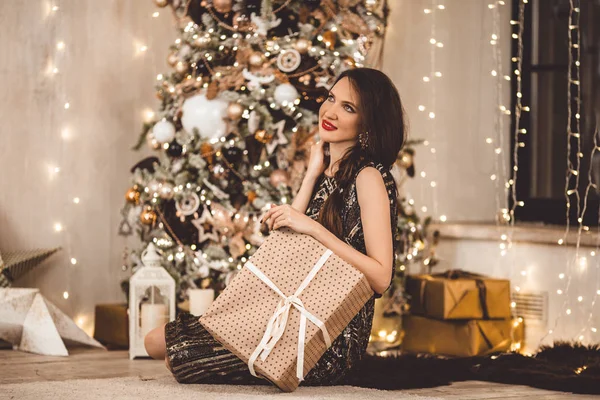 Schöne junge Frau trägt Luxuskleid vor dem Hintergrund eines Weihnachtsbaums mit Geschenkschachtel — Stockfoto