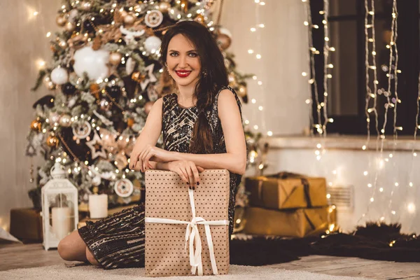 Όμορφη νεαρή γυναίκα φοράει φόρεμα πολυτελείας φόντο ένα χριστουγεννιάτικο δέντρο με το κιβώτιο δώρων — Φωτογραφία Αρχείου