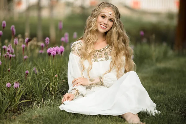 Feliz sonrisa chica rubia está sentado en la hierba en el parque de primavera con flores de color púrpura —  Fotos de Stock