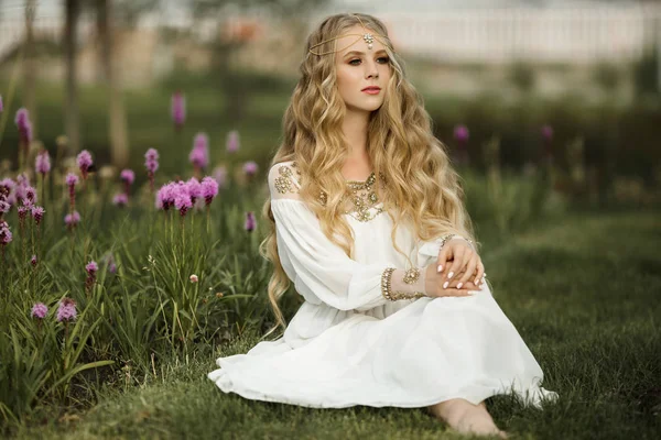 Feliz chica rubia sonriente con el pelo largo está sentado en la hierba en el parque de primavera con flores púrpuras — Foto de Stock