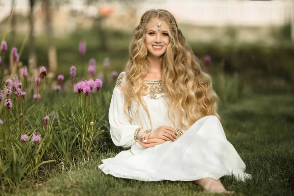 Feliz chica rubia sonriente con el pelo largo está sentado en la hierba en el parque de primavera con flores púrpuras —  Fotos de Stock