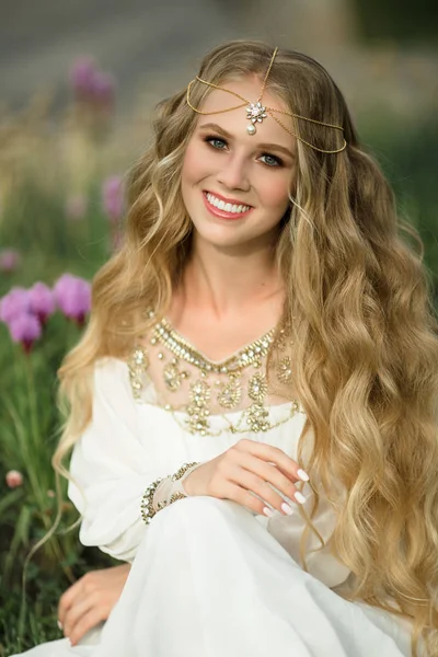Porträtt av glada leende blond flicka med långt hår sitter på gräset i spring park med lila blommor — Stockfoto